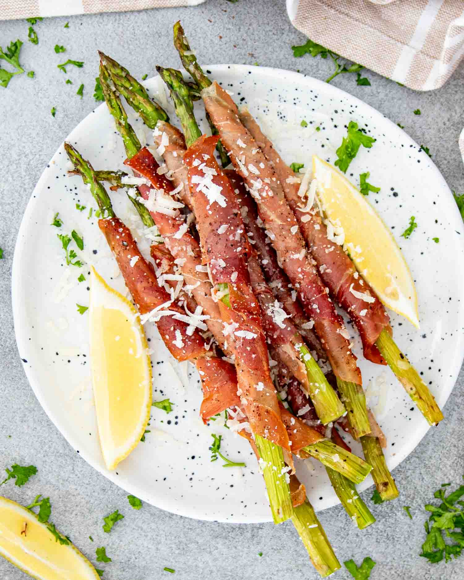 a serving of prosciutto wrapped asparagus on a white plate garnished with parmesan cheese and lemon wedges.