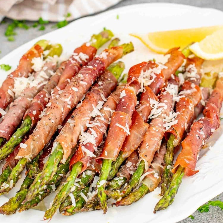 prosciutto wrapped asparagus on a serving platter garnished with parmesan cheese and lemon wedges.
