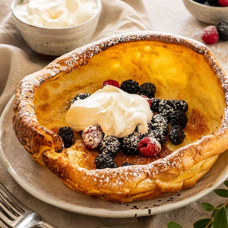 A golden Dutch baby pancake served on a white plate topped with fresh mixed berries and whipped cream.