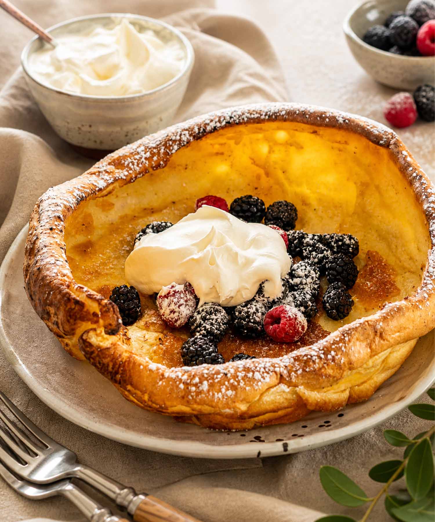 A golden Dutch baby pancake served on a white plate topped with fresh mixed berries and whipped cream.