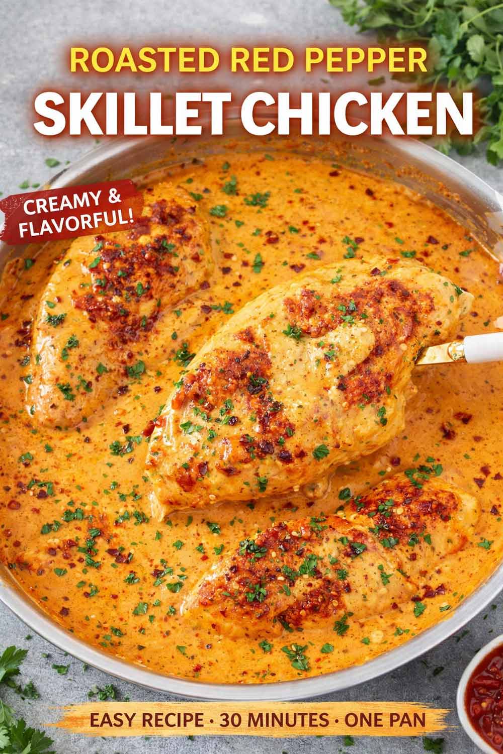A top down view of a silver skillet filled with three golden seared chicken breasts simmering in a vibrant creamy orange roasted red pepper sauce topped with fresh parsley and red pepper flakes.