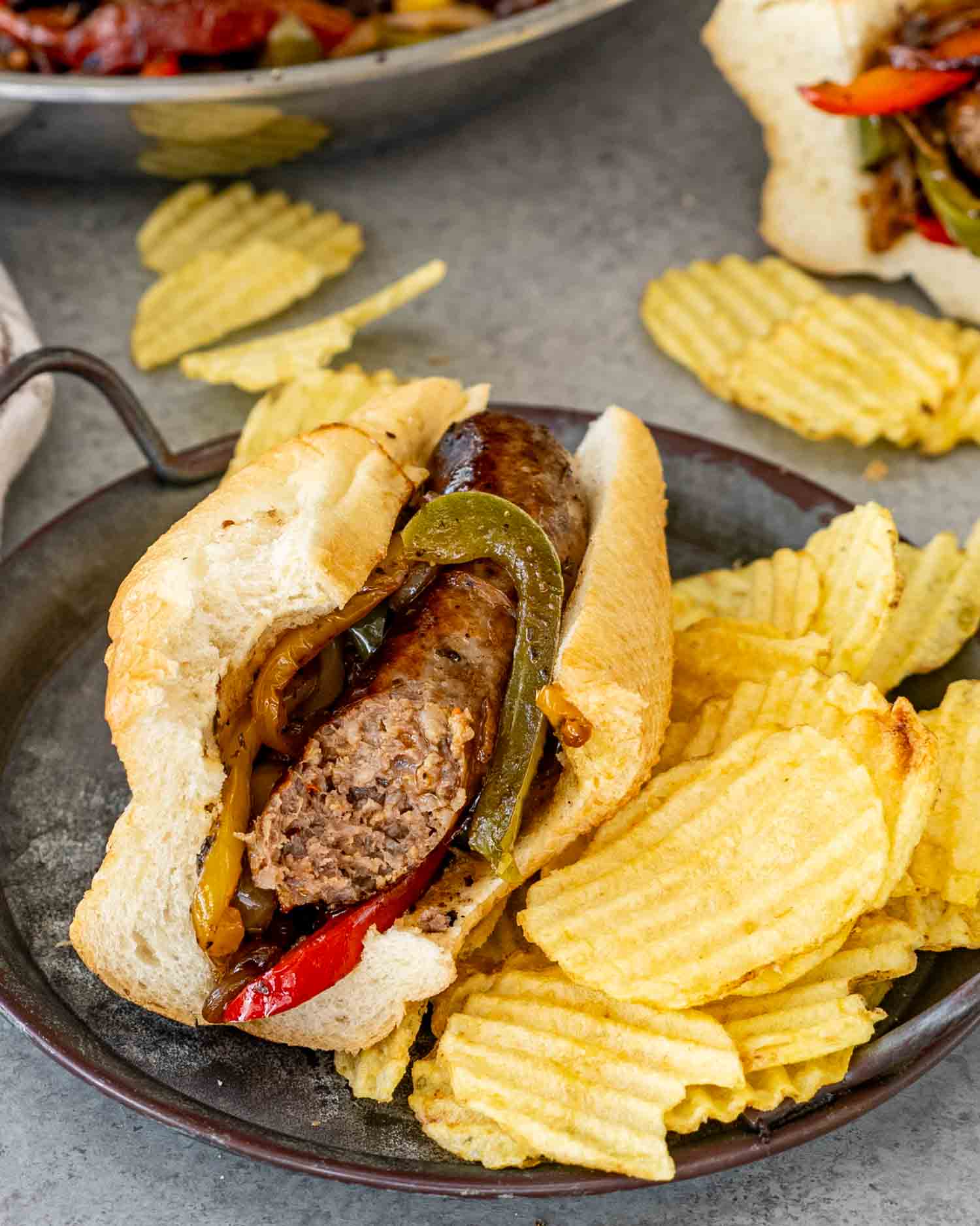 sausage peppers and onions in a hoagie roll alongside some potato chips.