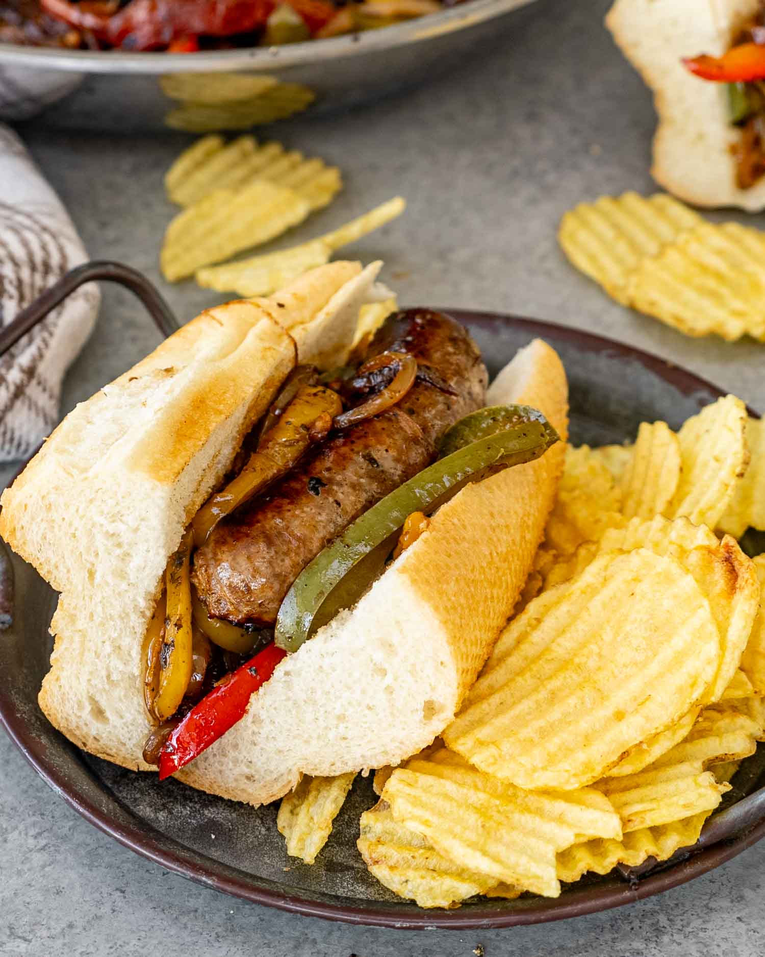 sausage peppers and onions in a hoagie roll alongside some potato chips.