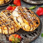 two banana nutella stuffed waffles on a nice metal plate drizzled with chocolate syrup and powdered sugar, along some sliced strawberries.