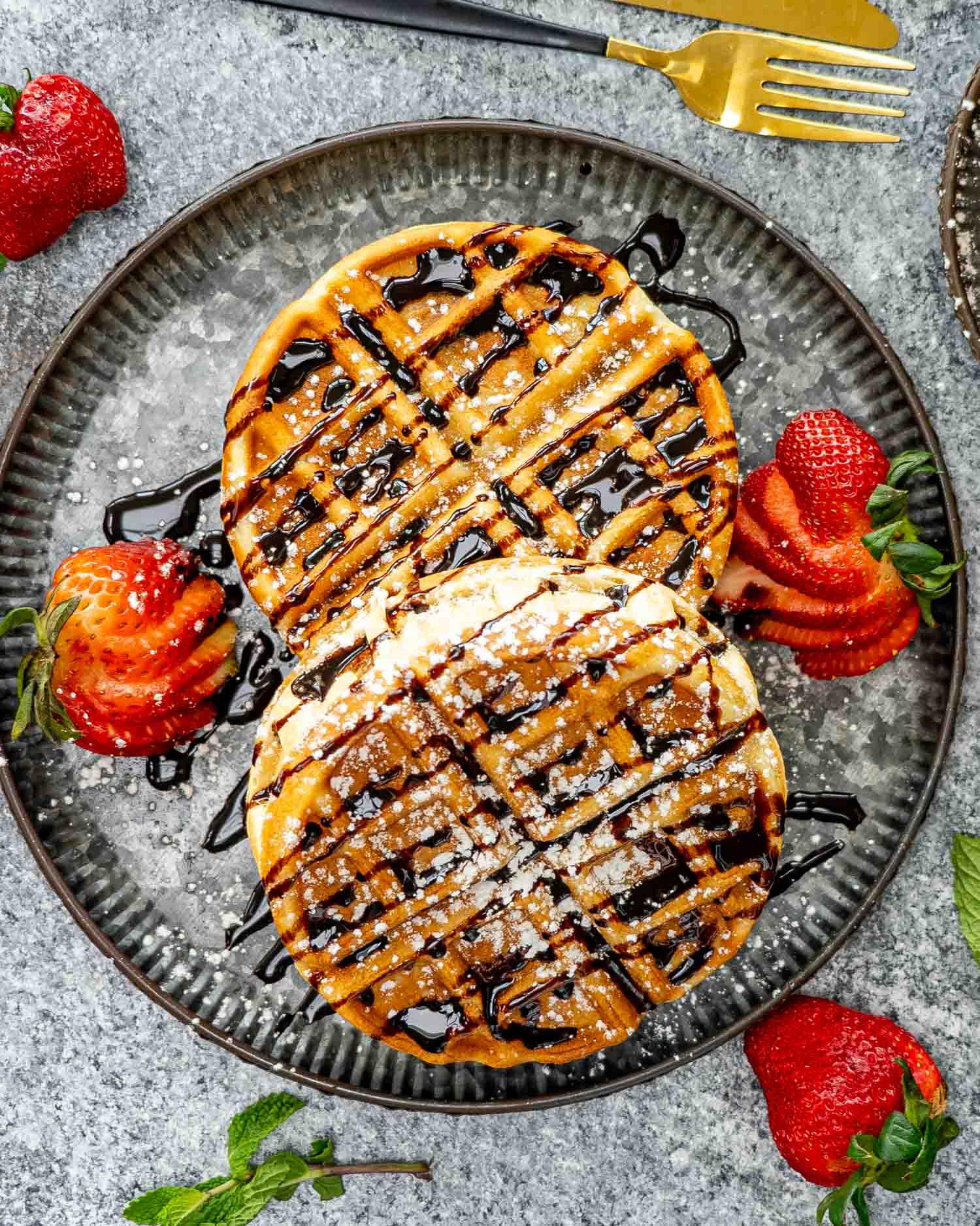 two banana nutella stuffed waffles on a nice metal plate drizzled with chocolate syrup and powdered sugar, along some sliced strawberries.