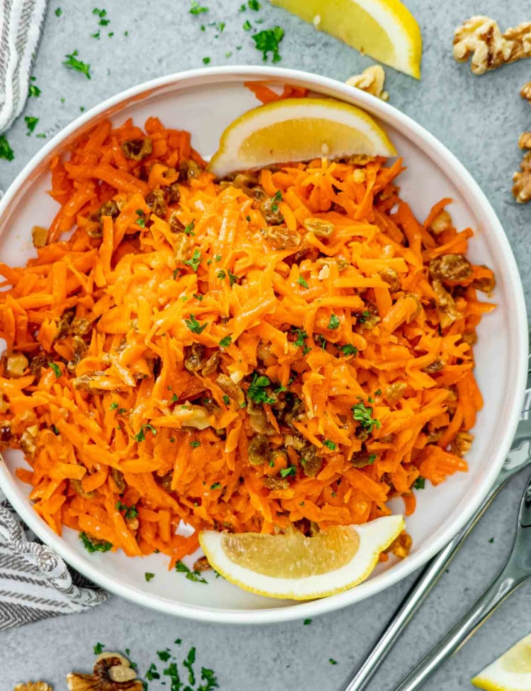 fresh carrot raisin salad in a white bowl garnished with parsley and lemon wedges.