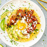 A vibrant plate of Huevos Rancheros featuring a sunny-side-up egg on a corn tortilla topped with refried beans, ranchero sauce, crumbled cheese, and avocado slices, garnished with fresh cilantro.