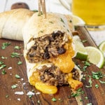 a beef burrito cut in half with melty cheese, on a cutting board with a glass of beer in the background.