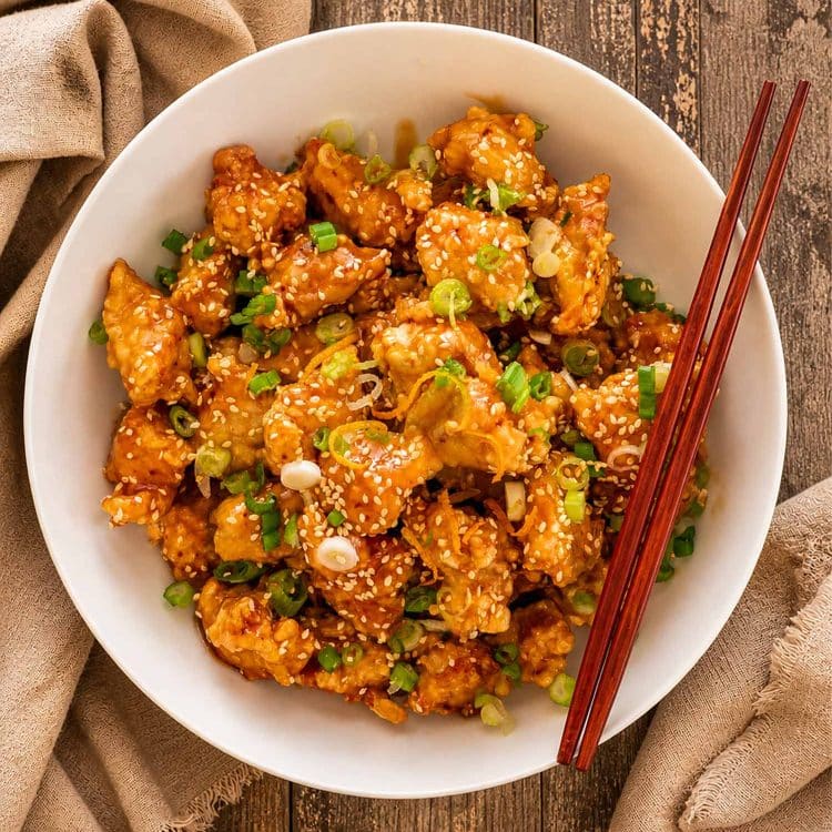 A bowl of crispy orange chicken topped with sesame seeds and finely chopped green onions. The chicken pieces are golden and glossy, coated in a rich orange sauce.