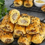 A plate stacked with golden brown pork and apple sausage rolls, topped with black and white sesame seeds. One sausage roll is cut in half, revealing the juicy sausage filling inside the flaky puff pastry. A cooling rack with more sausage rolls and a bowl of mustard are visible in the background.