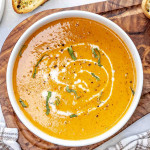 a white bowl filled with roasted red pepper soup drizzled with some cream and garnished with fresh basil.