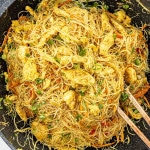 A vibrant dish of Singapore noodles served in a large black wok. The thin rice vermicelli noodles are mixed with chicken pieces, shrimp, and vegetables such as carrots, bell peppers, and green onions. The dish is seasoned with curry powder, giving it a yellowish hue, and garnished with fresh cilantro. Chopsticks are placed in the noodles, indicating it's ready to be served.