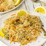 A plate of Filipino pancit with thin rice noodles, shredded vegetables, and chicken, garnished with lemon wedges and fresh herbs.