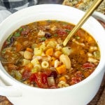 White bowl filled with pasta e fagioli soup, featuring beans, pasta, and vegetables, with garlic bread on the side.