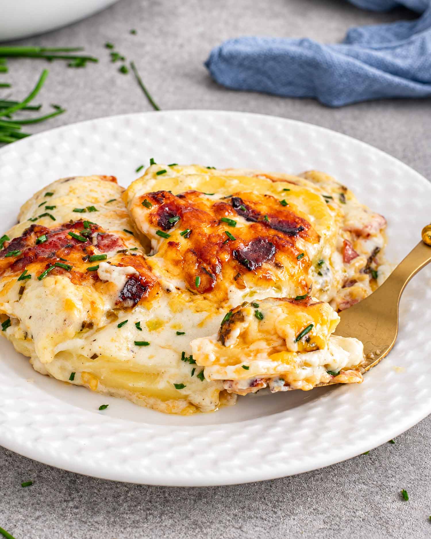 Loaded scalloped potatoes with creamy sauce, golden cheese, bacon, and chives on a white plate, with a forkful lifted.