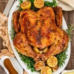 Overhead view of dry brined roast turkey surrounded by herbs, roasted lemons, and garlic, with gravy and mashed potatoes nearby.