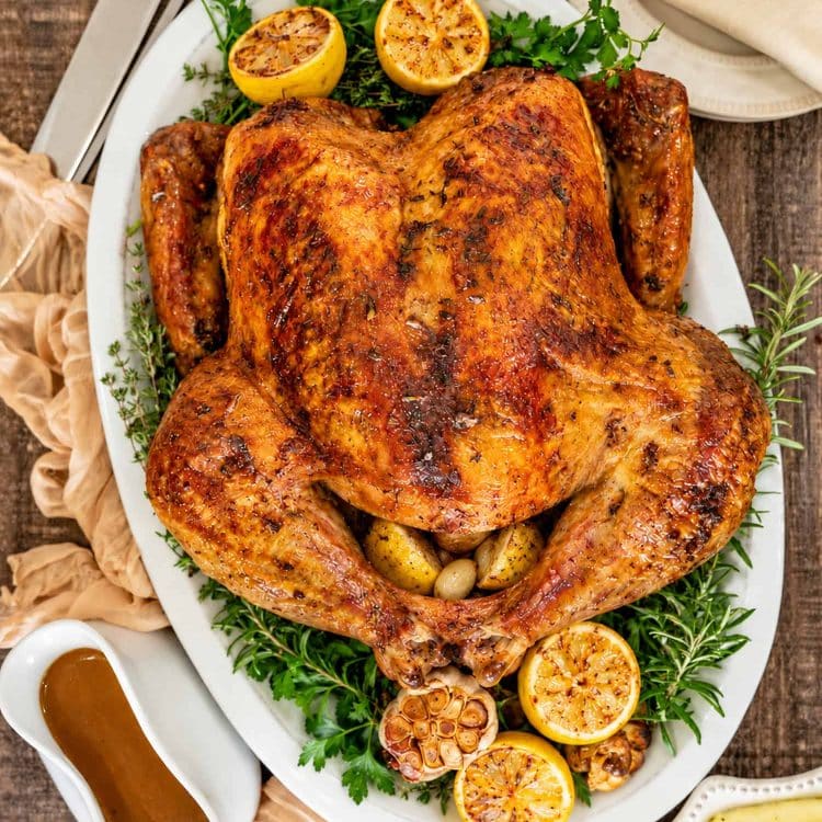 Overhead view of dry brined roast turkey surrounded by herbs, roasted lemons, and garlic, with gravy and mashed potatoes nearby.