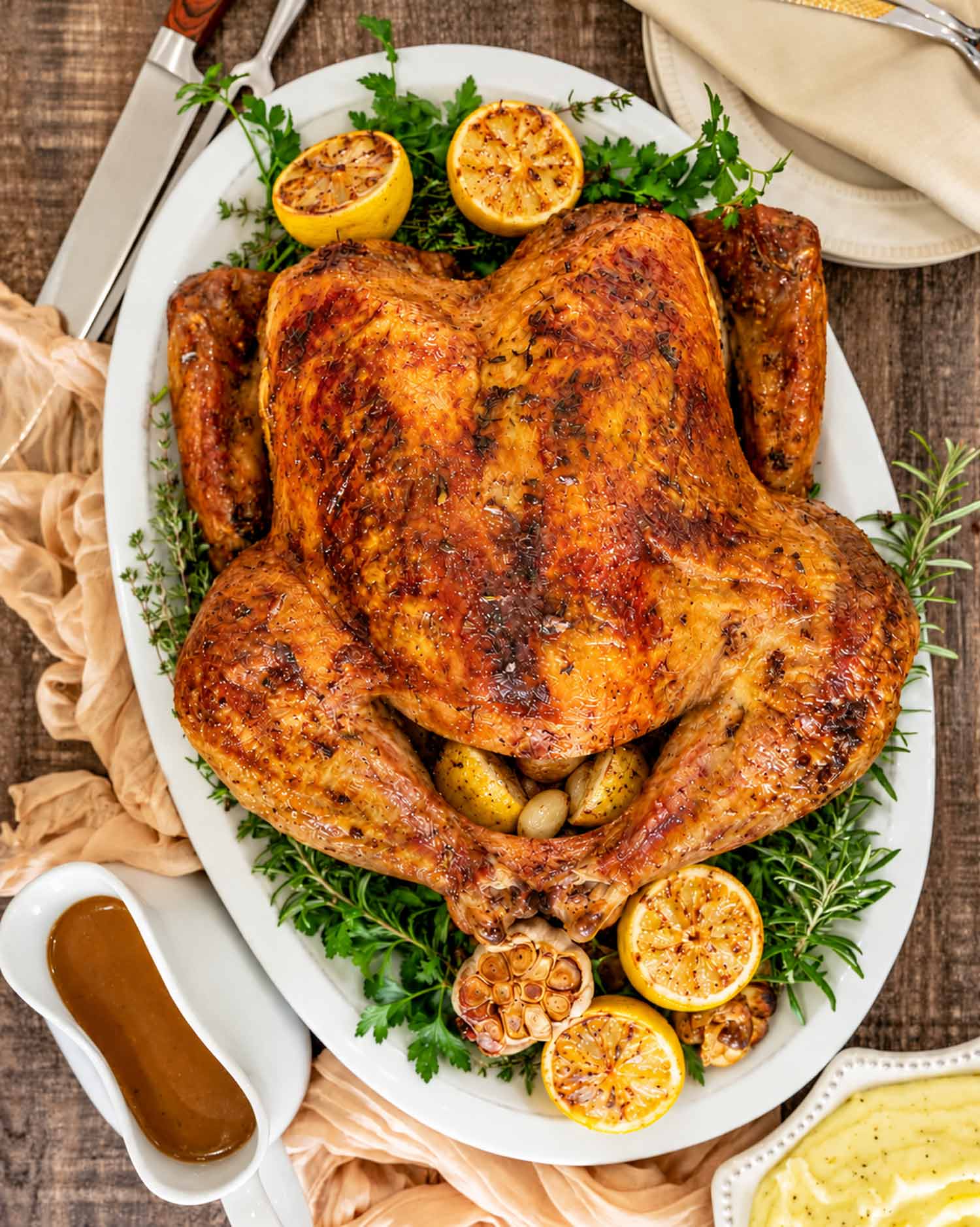 Overhead view of dry brined roast turkey surrounded by herbs, roasted lemons, and garlic, with gravy and mashed potatoes nearby.