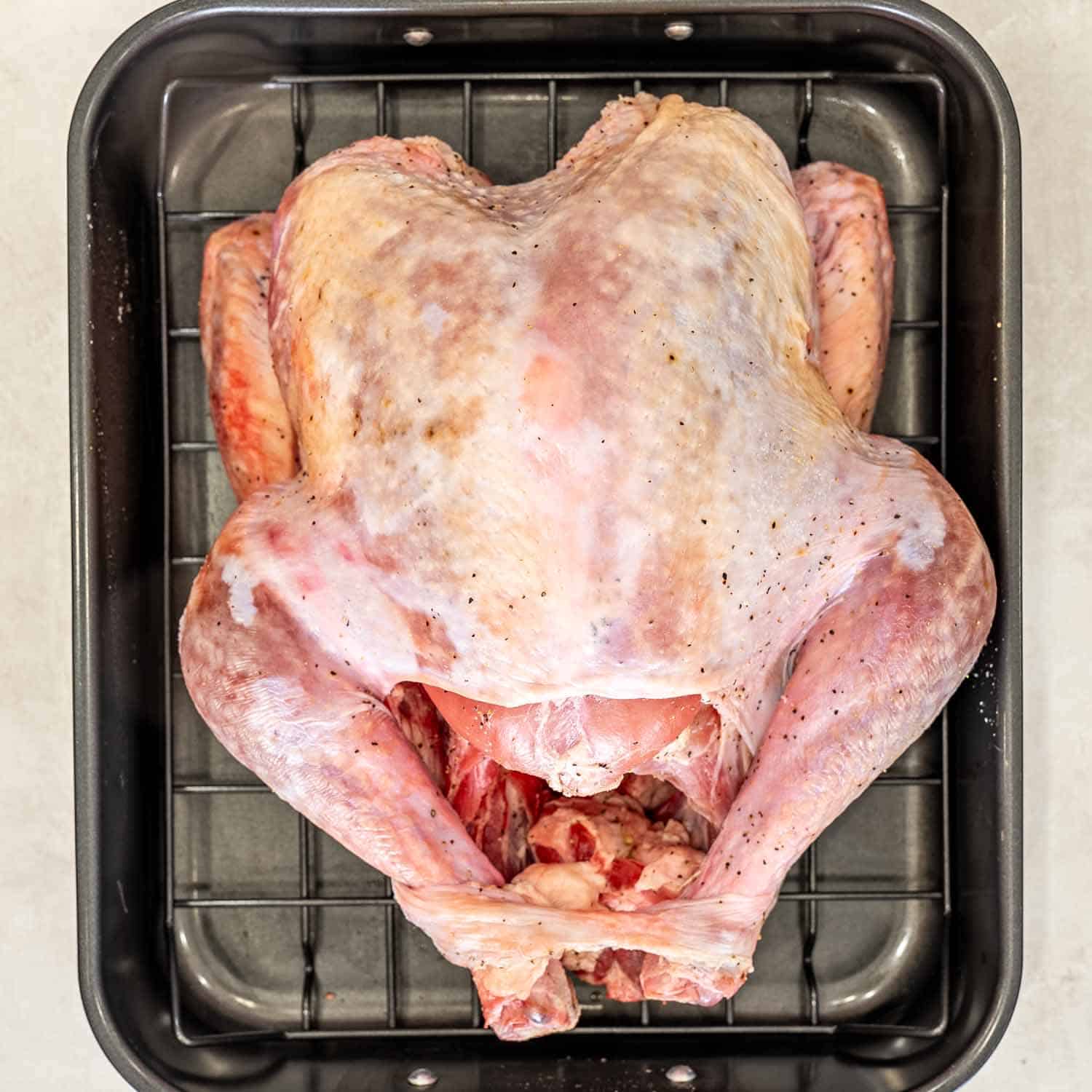 Overhead view of a dry brined raw turkey seasoned with salt and pepper, resting on a rack in a roasting pan.
