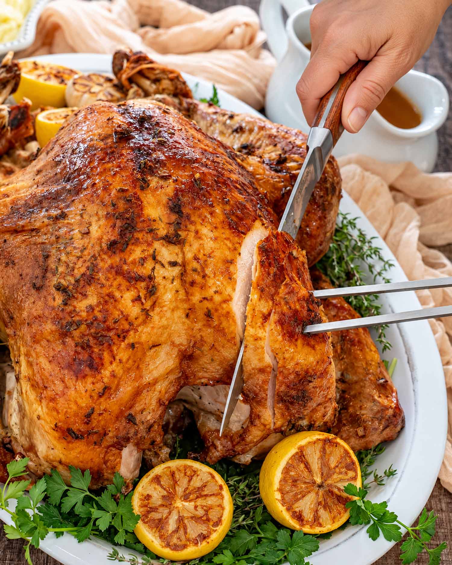 Hand carving golden roast turkey breast with a knife and carving fork, surrounded by roasted lemons and fresh herbs.