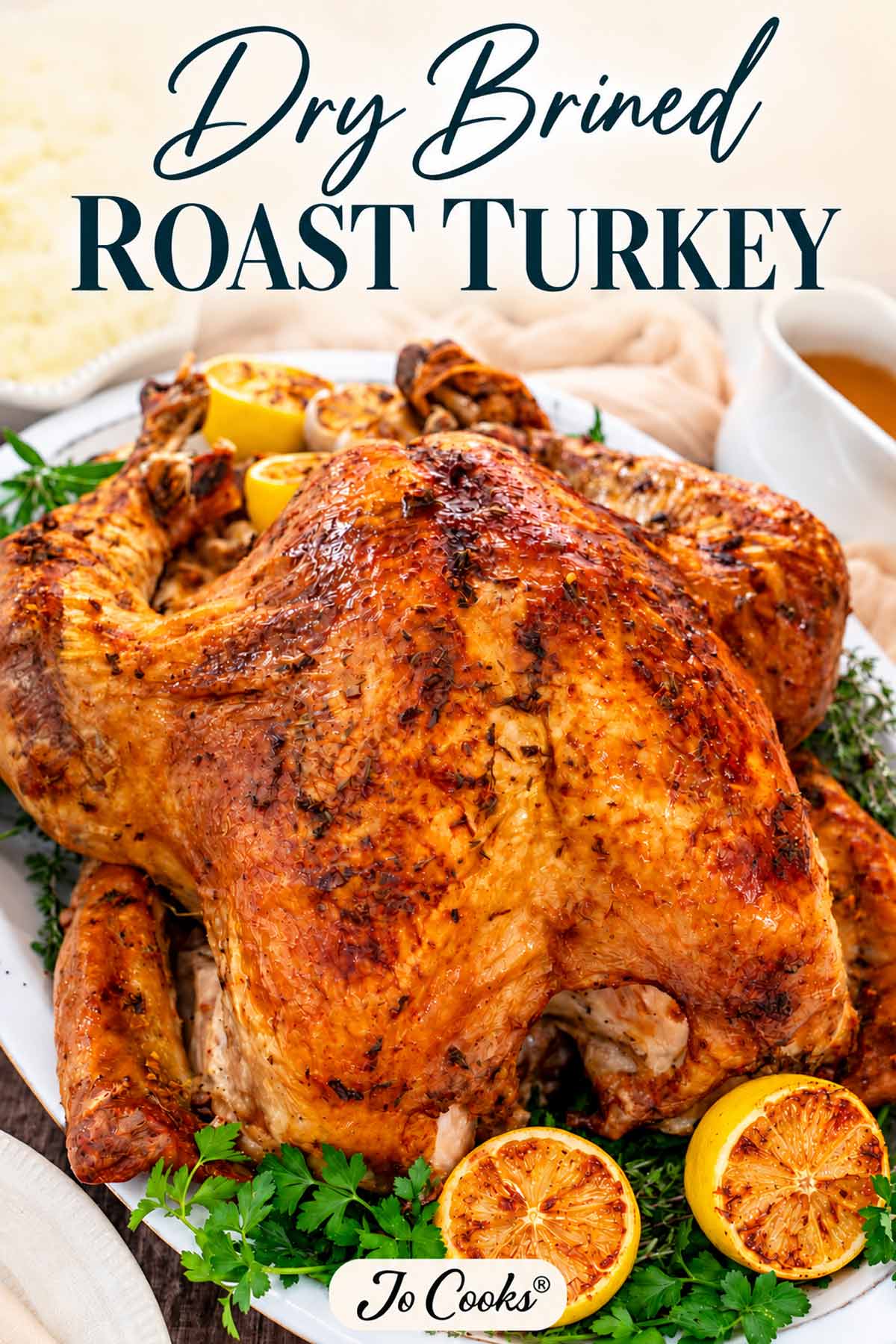 Golden roast turkey garnished with herbs and roasted lemons, with Dry Brined Roast Turkey title and Jo Cooks logo.