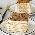 Two slices of Carlota de Café served on white plates with dark rims, showcasing the delicate layers of Maria cookies and creamy coffee filling. The top is dusted with cocoa and coffee, and the plates are garnished with a few scattered coffee grounds.