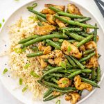 a serving of chicken green bean stir fry in a white plate with a serving of rice.