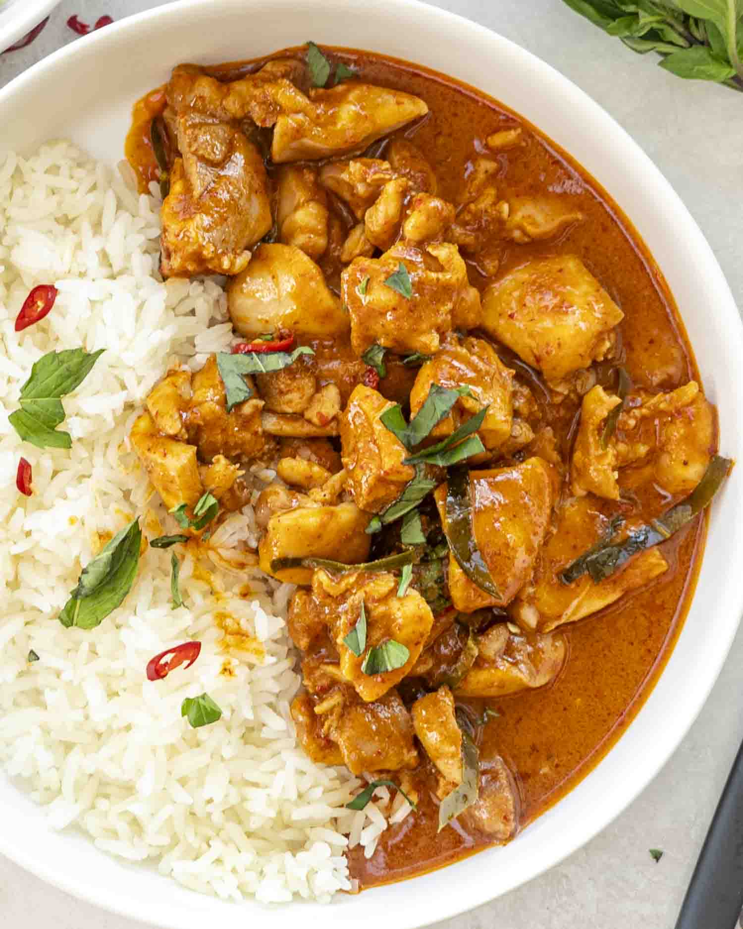 A bowl of Panang Chicken Curry served with fluffy jasmine rice, garnished with fresh basil and red chili slices, showcasing a rich, vibrant curry sauce.