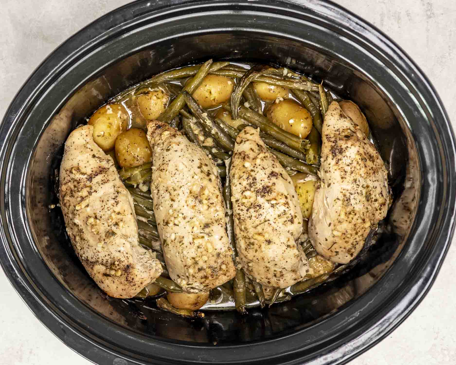 Close-up of slow-cooked chicken breasts, golden baby potatoes, and green beans, all seasoned with garlic, ranch, and butter, inside a black slow cooker.