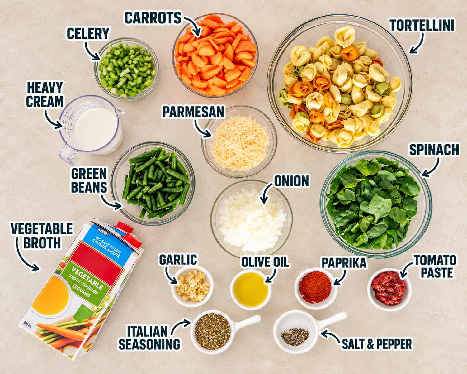 Labeled vegetable tortellini soup ingredients on beige stone, including tortellini, spinach, carrots, green beans, cream, Parmesan, broth and seasonings.