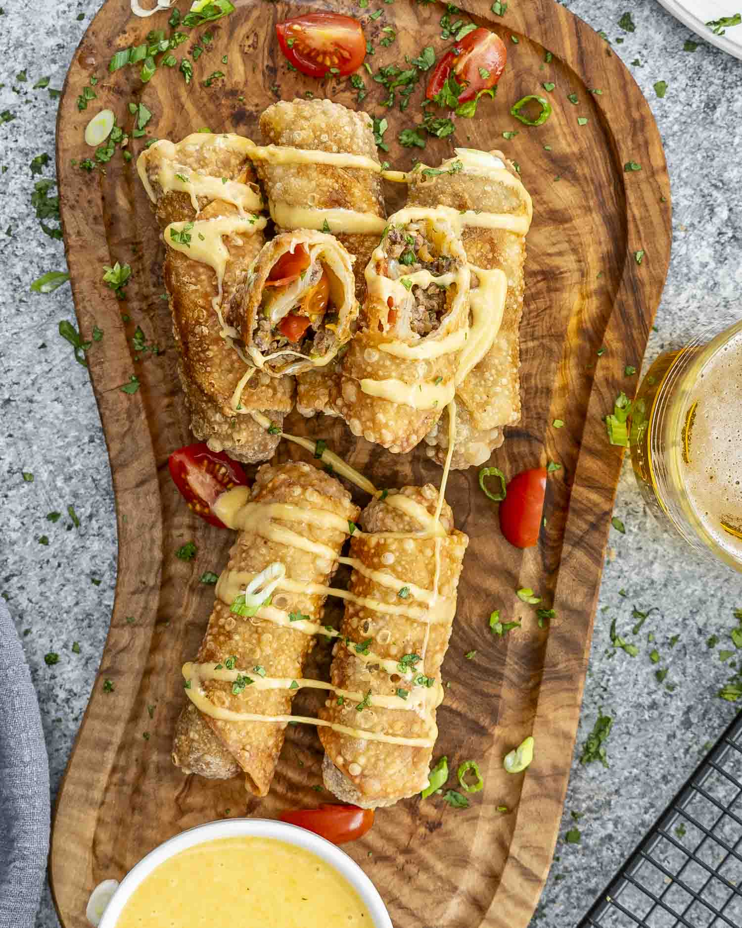 Crispy beef taco egg rolls drizzled with creamy beer cheese sauce, garnished with fresh cilantro and green onions on a wooden board.
