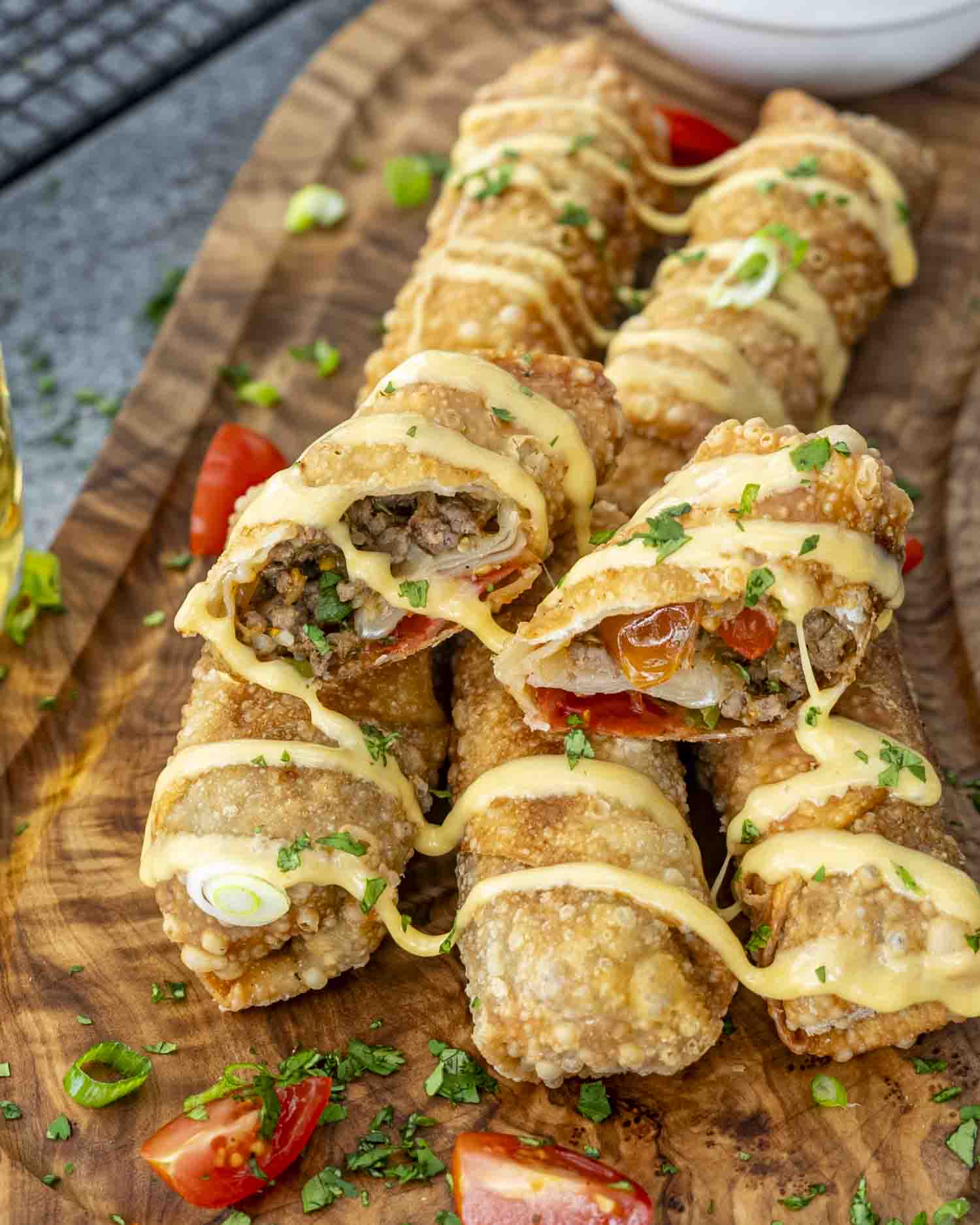 Crispy beef taco egg rolls drizzled with creamy beer cheese sauce, garnished with fresh cilantro and green onions on a wooden board.