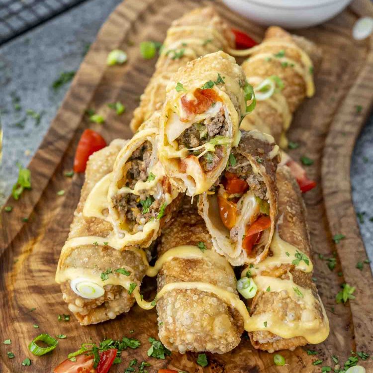 Crispy beef taco egg rolls drizzled with creamy beer cheese sauce, garnished with fresh cilantro and green onions on a wooden board.
