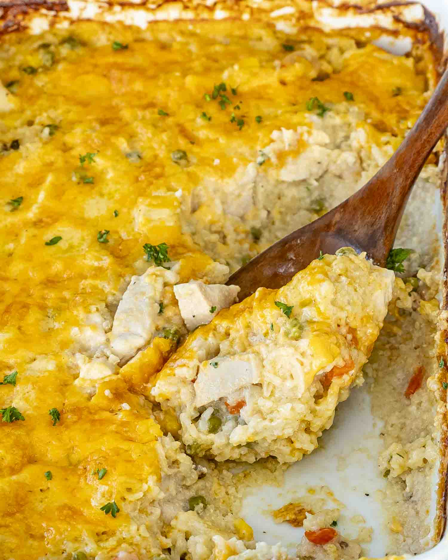A cheesy, golden-baked chicken and rice casserole in a white dish, with a wooden spoon scooping out a creamy serving.