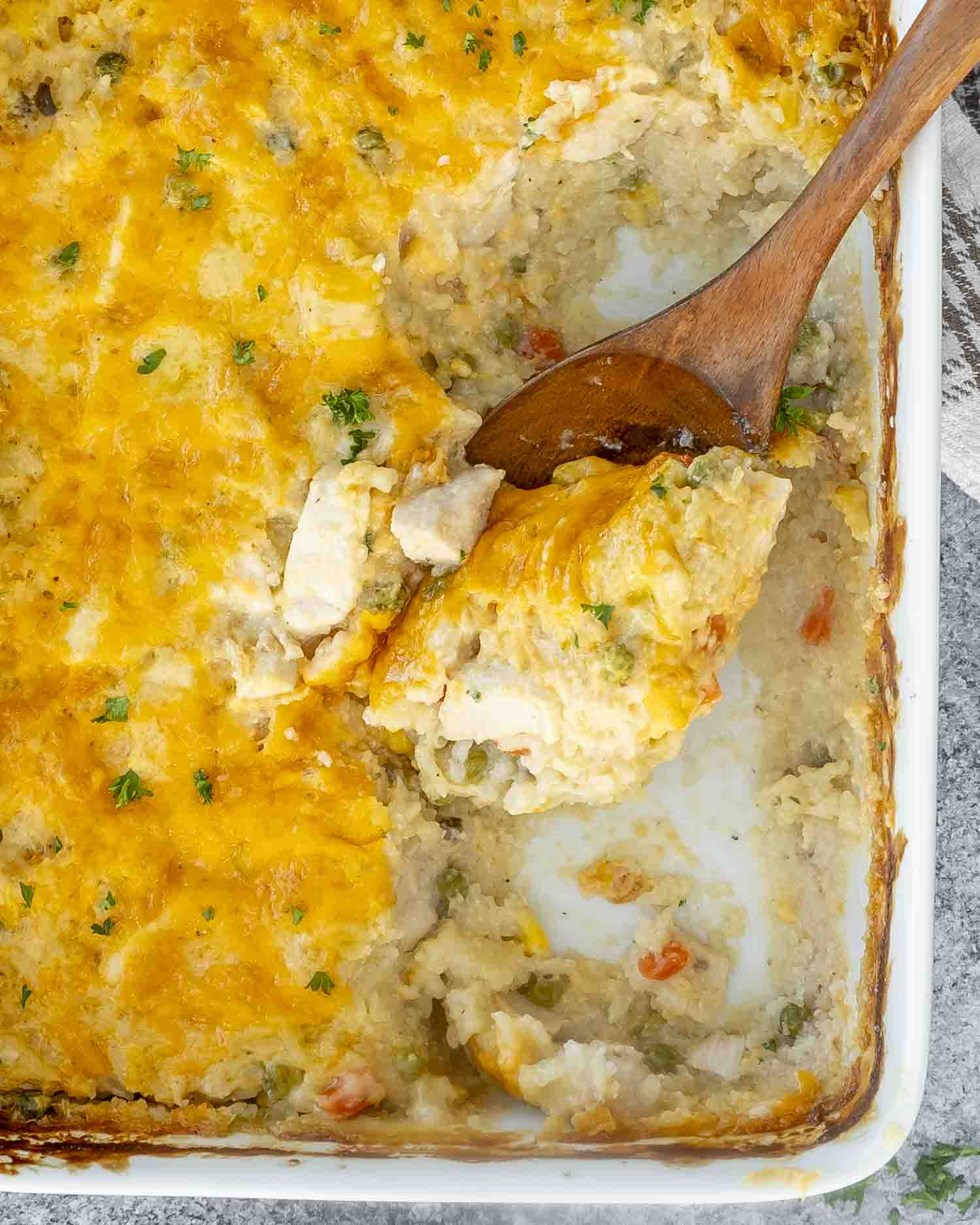 A cheesy, golden-baked chicken and rice casserole in a white dish, with a wooden spoon scooping out a creamy serving.