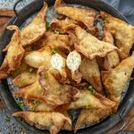 A black metal serving tray filled with crispy, golden-brown chicken fried wontons, garnished with finely chopped green onions. One wonton is cut open, revealing a juicy, well-seasoned chicken and cabbage filling. A small bowl of dipping sauce with chili flakes sits in the background, adding a rich, savory contrast to the crunchy wontons.
