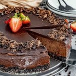 A rich and creamy chocolate cheesecake with a glossy ganache topping, garnished with fresh strawberries and chocolate shavings. A slice is being lifted, revealing the velvety chocolate filling and a dark, crumbly chocolate cookie crust.