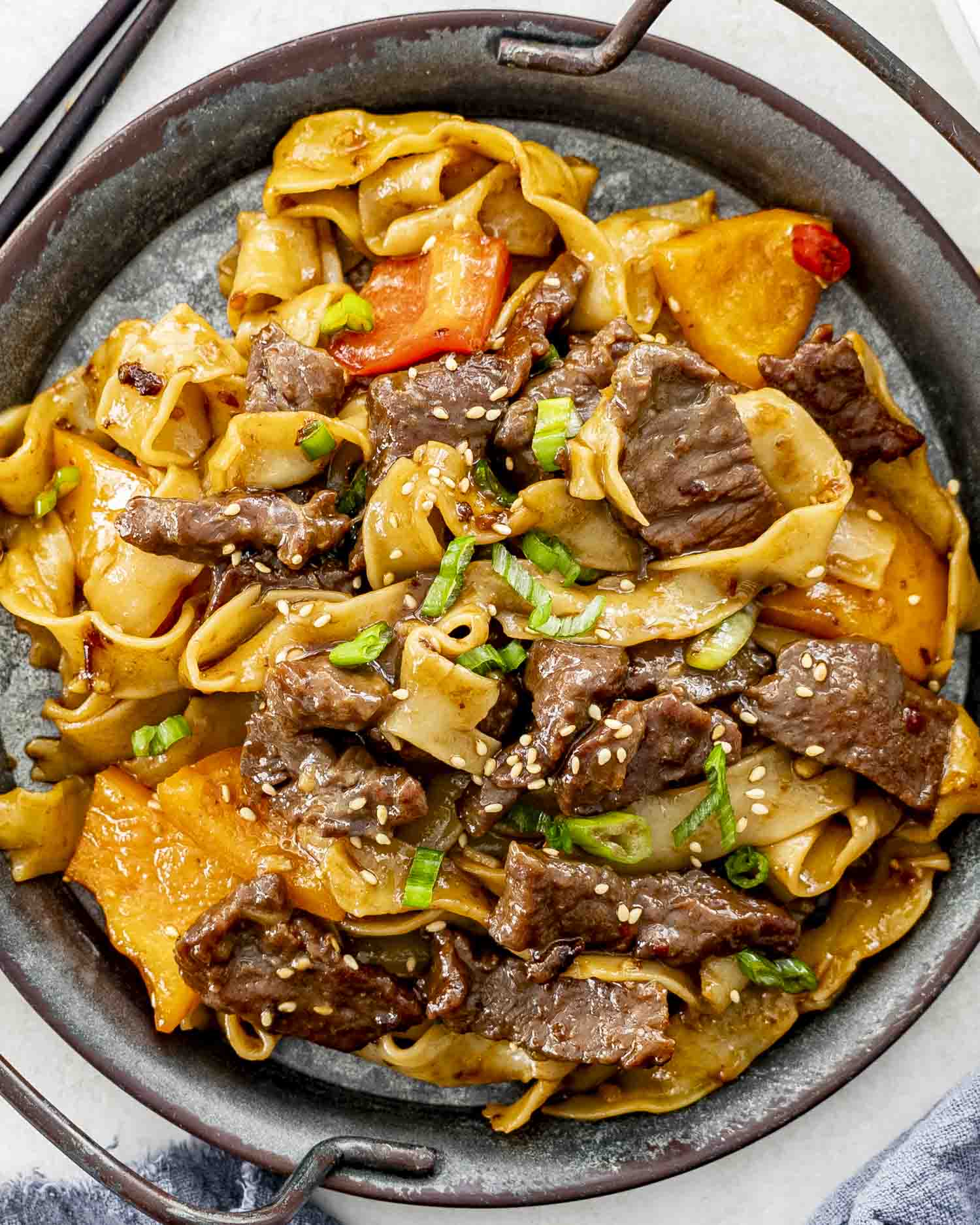 A serving of spicy beef noodles with tender beef strips, flat noodles, and bell peppers, garnished with sesame seeds and green onions.
