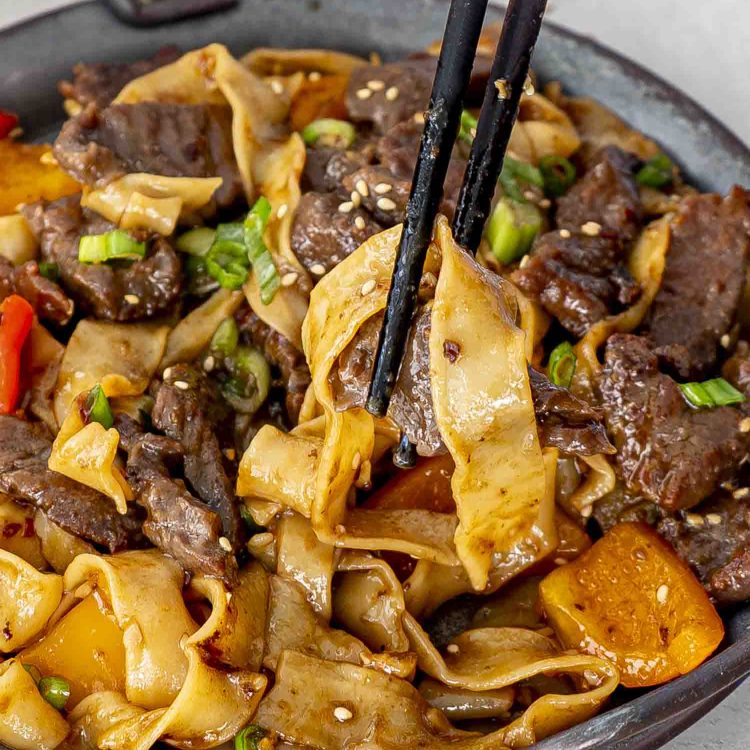 A close-up of spicy beef noodles with tender beef strips and flat noodles, lifted with chopsticks, garnished with green onions and sesame seeds.