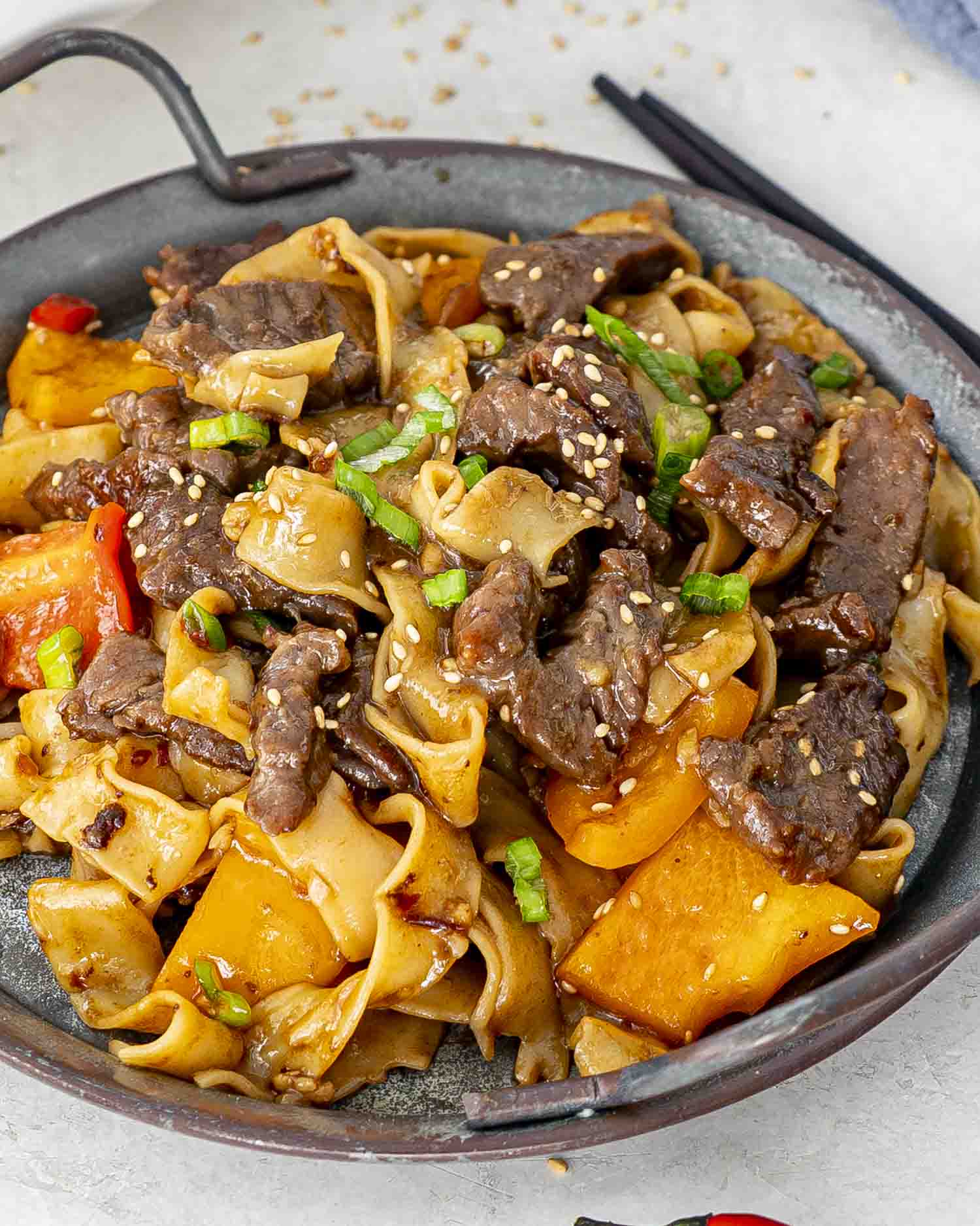 A serving of spicy beef noodles with tender beef strips, flat noodles, and bell peppers, garnished with sesame seeds and green onions.