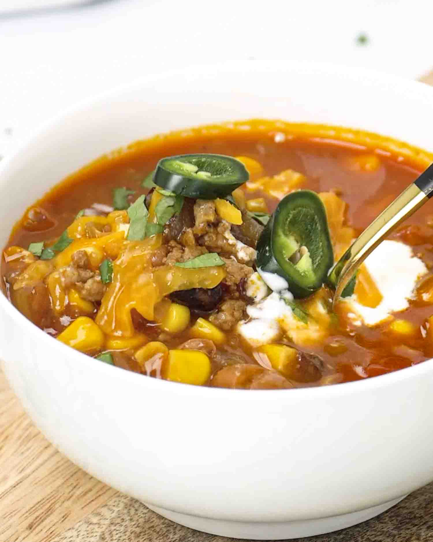 A close-up of taco soup in a white bowl, topped with melted cheese, sour cream, sliced jalapeños, and fresh cilantro, with a spoon scooping in.