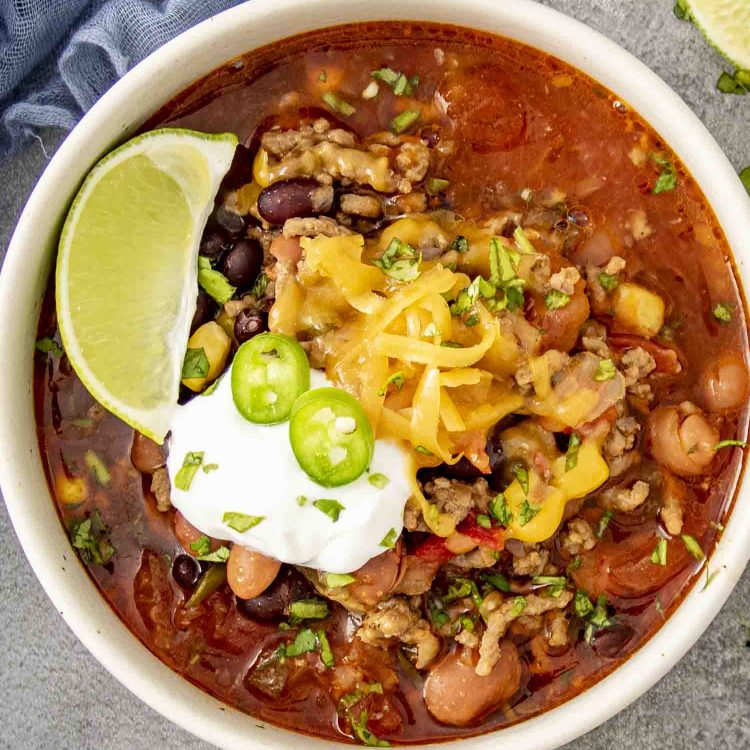 A bowl of taco soup topped with melted cheese, sour cream, sliced jalapeños, and fresh cilantro, served with a lime wedge on the side.