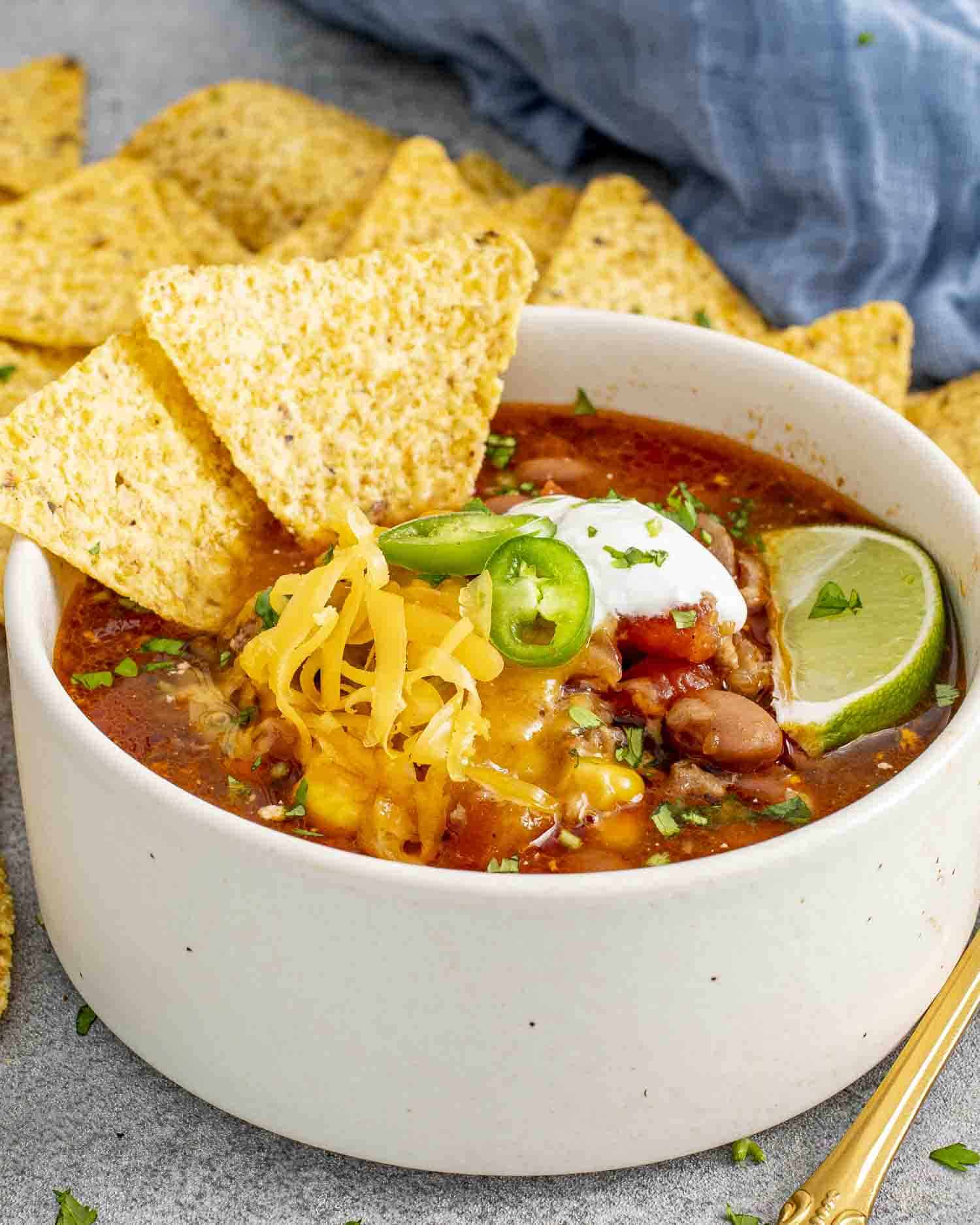 A bowl of taco soup topped with shredded cheese, sour cream, sliced jalapeños, and a lime wedge, served with tortilla chips on the side.