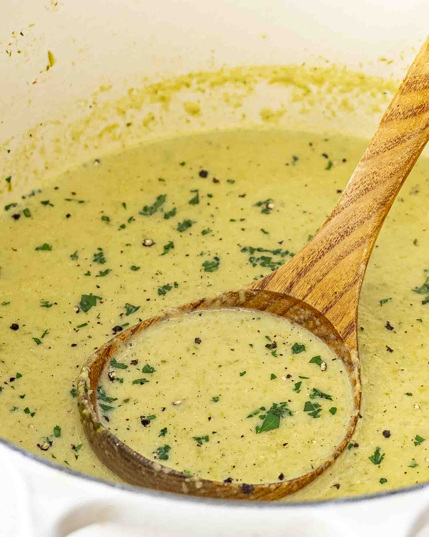freshly made asparagus soup in a beige dutch oven with a wooden ladle inside and garnished with parsley.