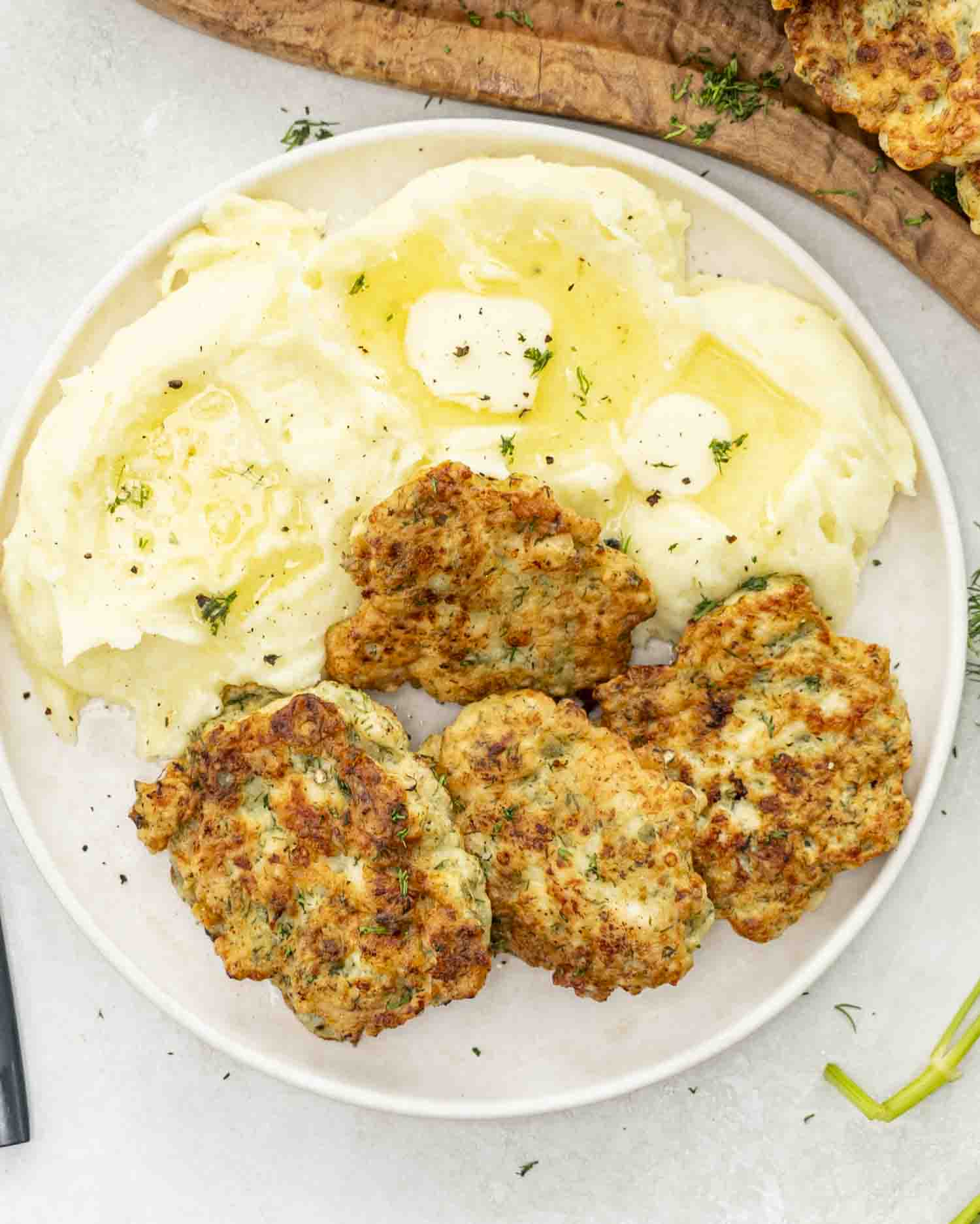 Crispy chicken fritters served with buttery mashed potatoes and fresh dill on a white plate—comfort food at its best.