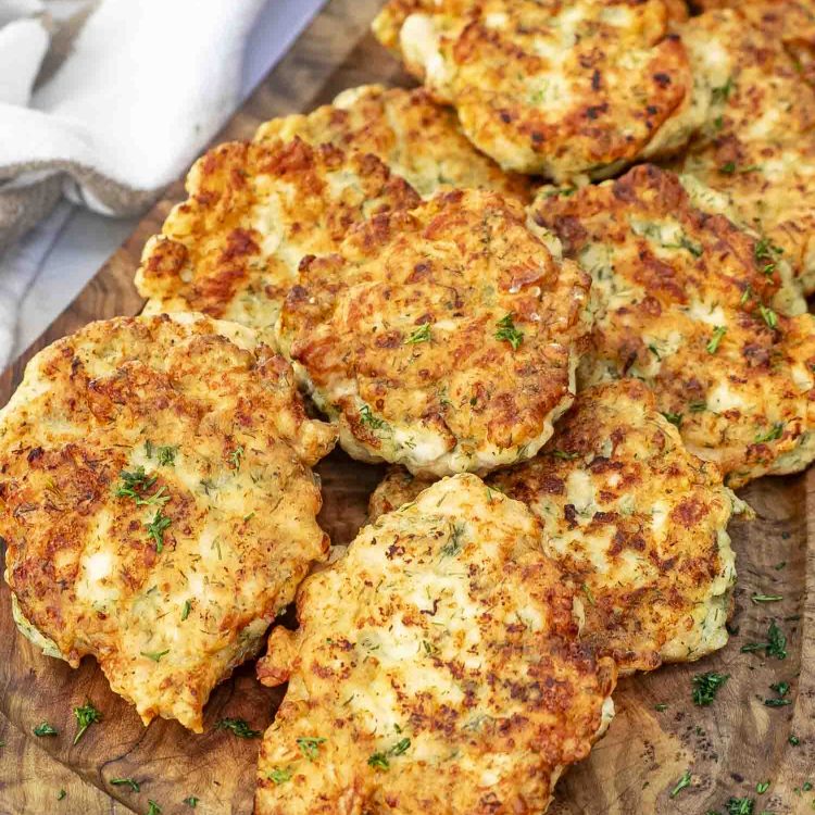 Golden chicken fritters on a wooden board, garnished with fresh dill and served with a bowl of creamy dipping sauce.