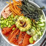 A sushi bowl filled with sushi rice topped with sliced salmon, avocado drizzled with soy sauce, cucumber, shredded carrots, edamame, and thin strips of nori.