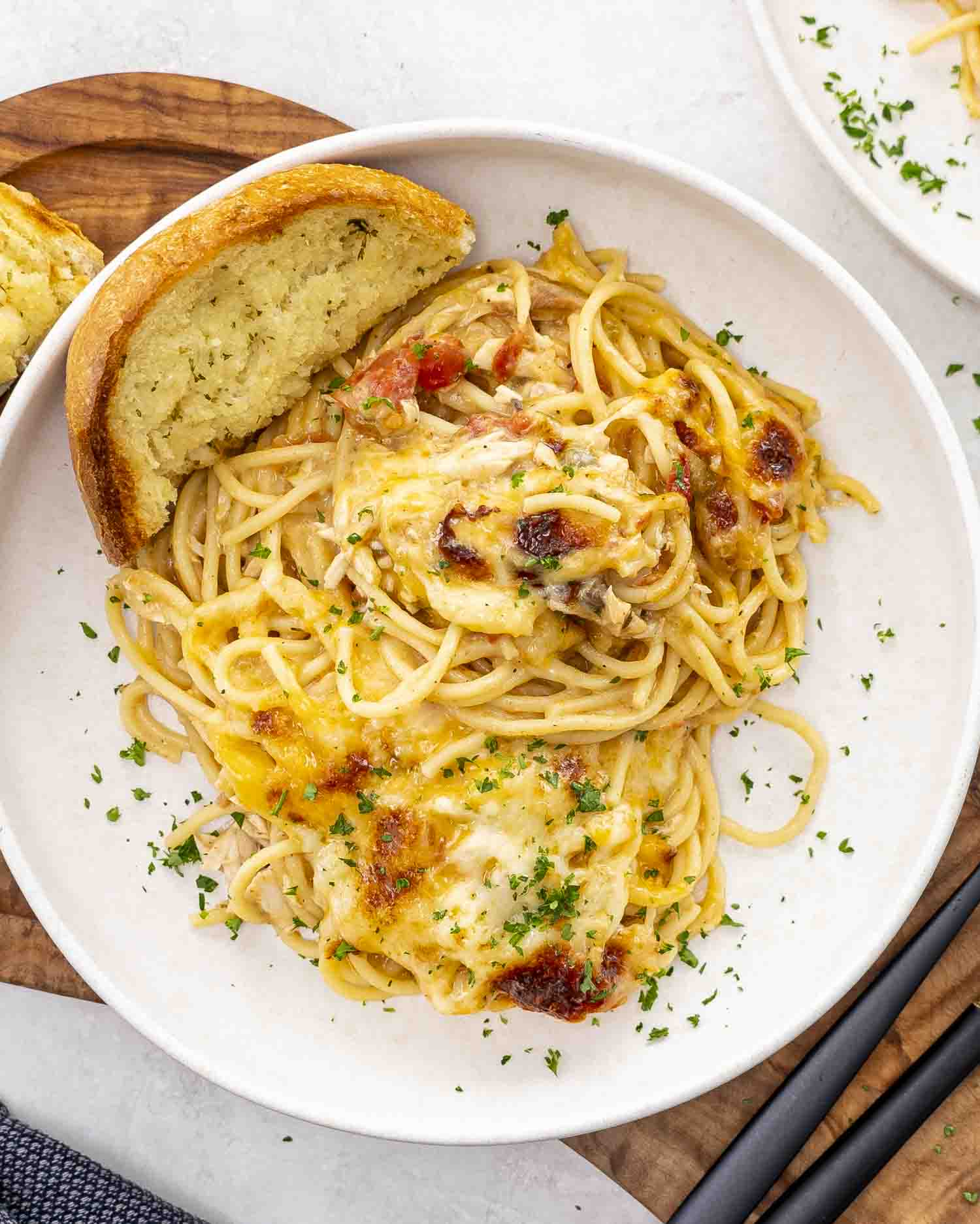 Creamy chicken spaghetti served on a white plate, topped with melted cheese and fresh parsley, paired with a slice of garlic bread.