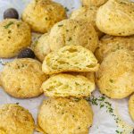 Golden brown gougères topped with herbs and grated cheese, arranged on parchment paper with whole nutmegs scattered around.