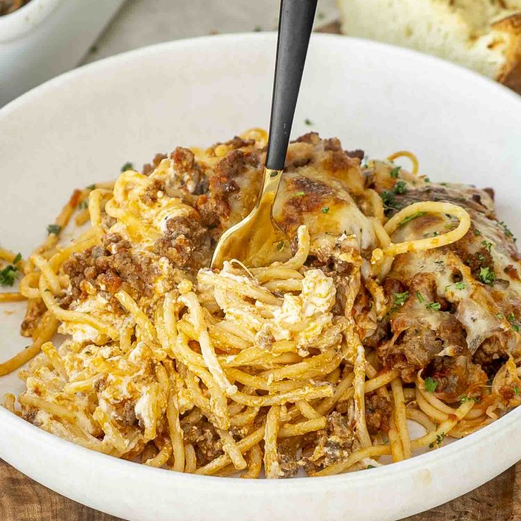 Close-up of creamy, cheesy baked spaghetti being twirled with a gold fork, showing layers of sauce, noodles, and melted mozzarella.